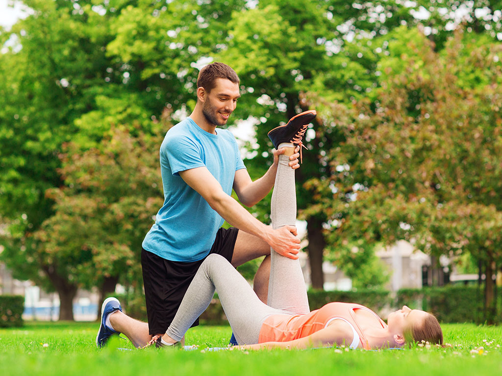 Male personal trainer assisting a woman with a hamstring stretch outdoors